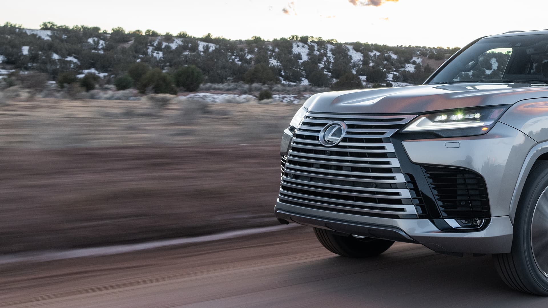 Silver Lexus LX being driven on a road next to a sandy terrain.