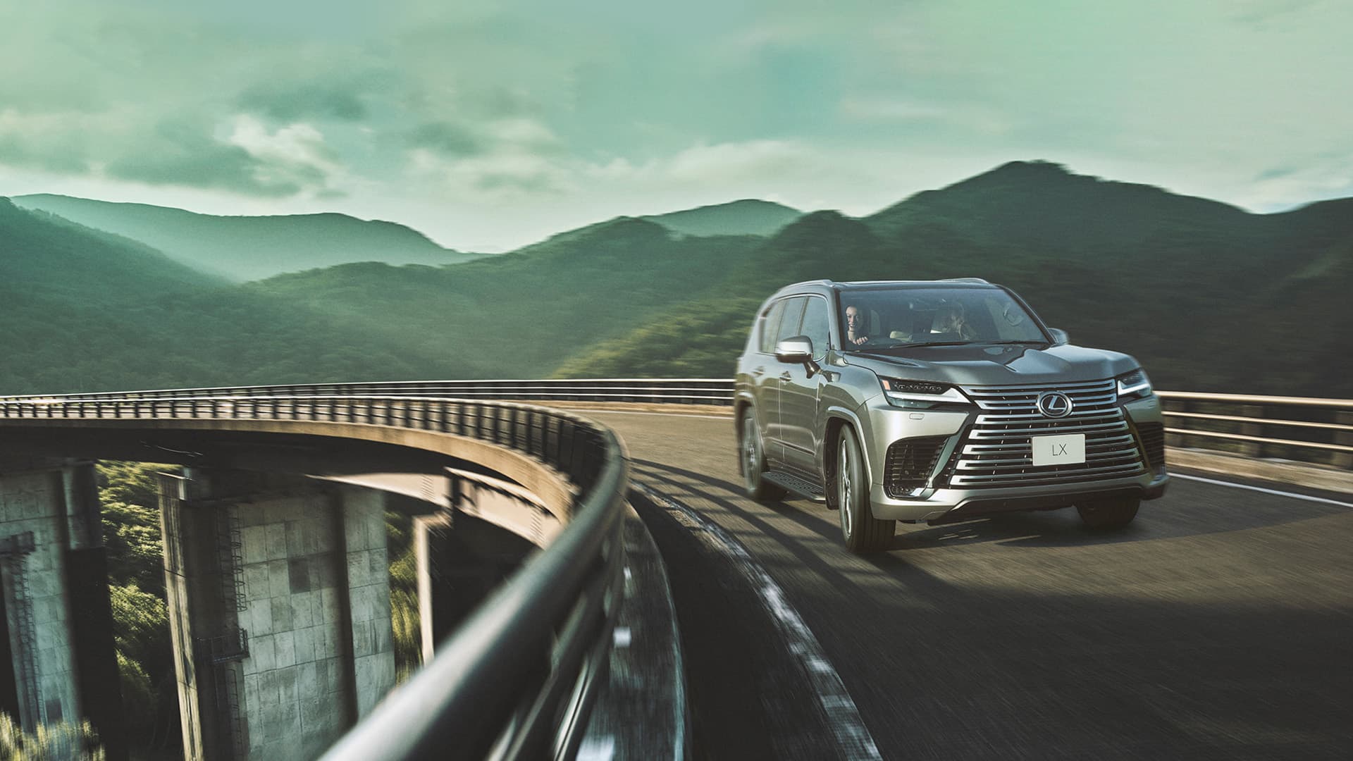 Silver Lexus LX being driven on a bridge with mountains in the backdrop.